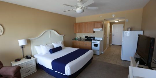 Accessible King Studio. Bed along left wall. Kitchen in the center of image. Dresser with television on the right side of the room.