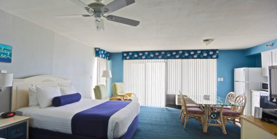 Spacious Queen Studio. Two queen beds on left. Large patio doors with blinds in front in the center of image. Dining room table with the kitchen on the right.