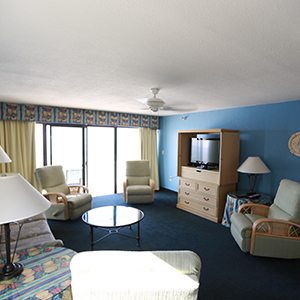 Living room. Two end tables with lamps flanking a sofa on the left. large patio door to balcony in the middle. Dresser with TV on top on the right. Three recliners visible around the room.